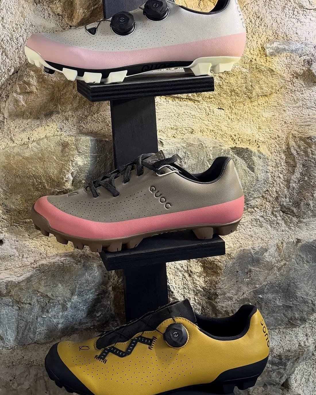 Trois paires de chaussures de vélo gravel sur un présentoir, devant un mur en pierre