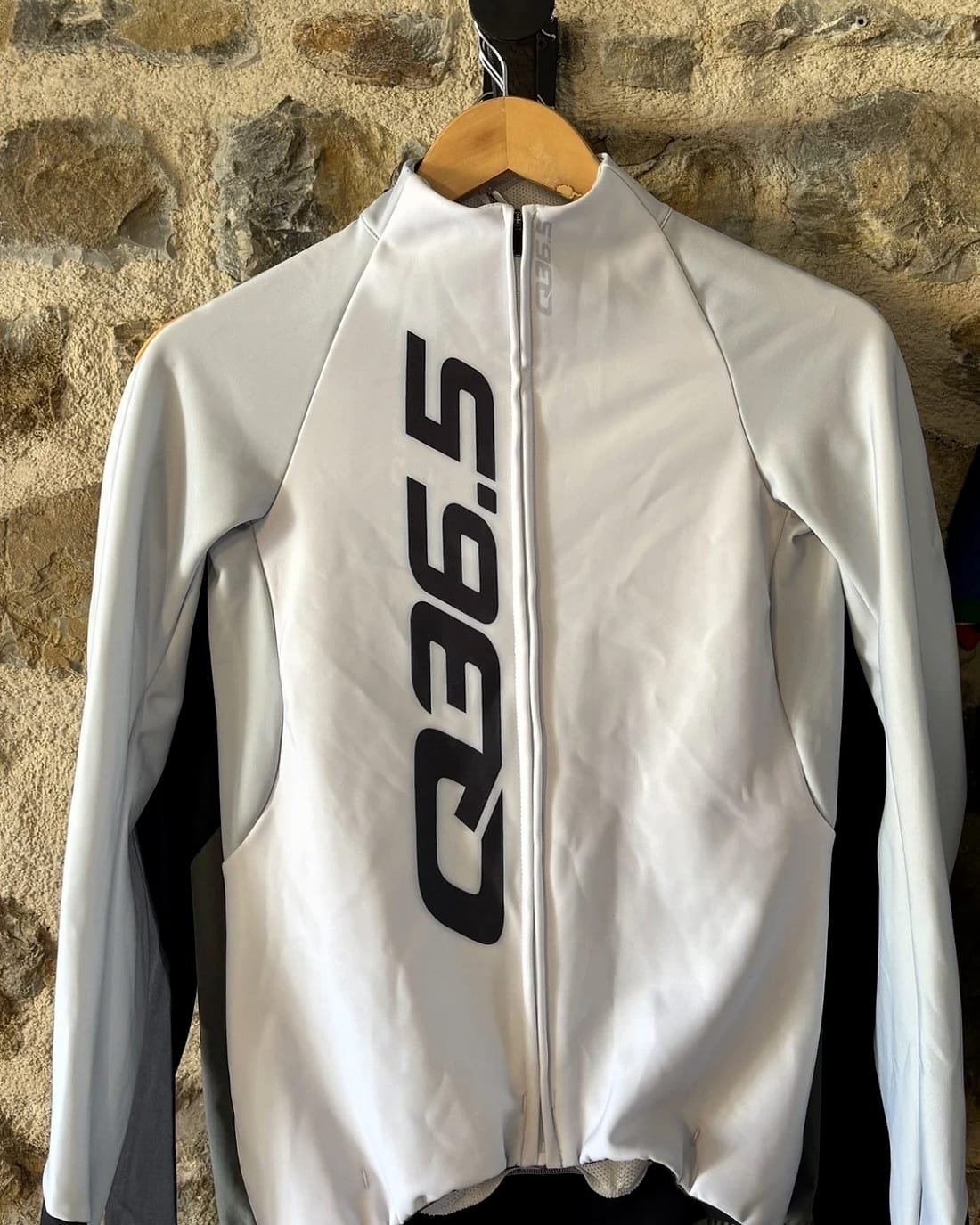 Veste de vélo Q36.5 blanche sur un cintre, devant un mur en pierre